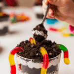 Child's hand with a spoon reaching for an individual serving of Oreo Dirt Cake with gummy worms.
