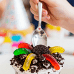 Child's hand with a spoon reaching for an individual serving of Oreo Dirt Cake with gummy worms.
