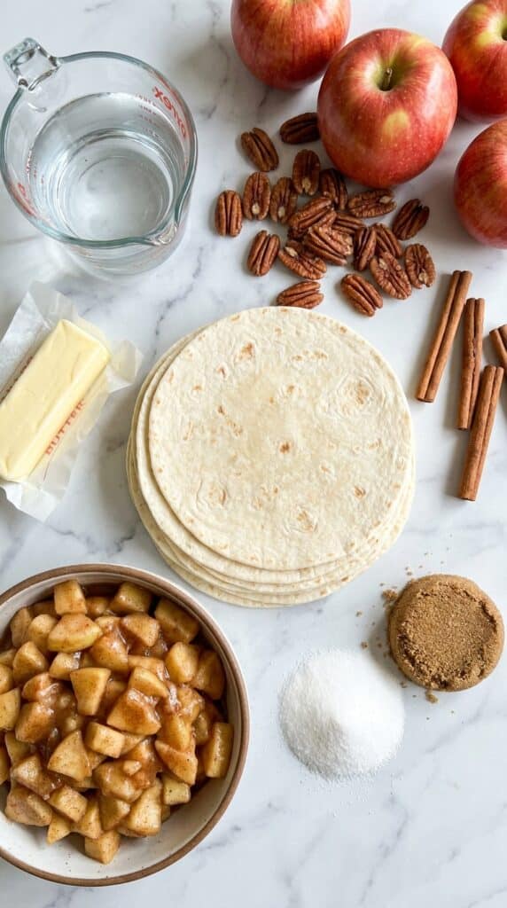 A flat lay showing flour tortillas, apple filling, butter, sugars, and pecans neatly arranged on a marble surface.