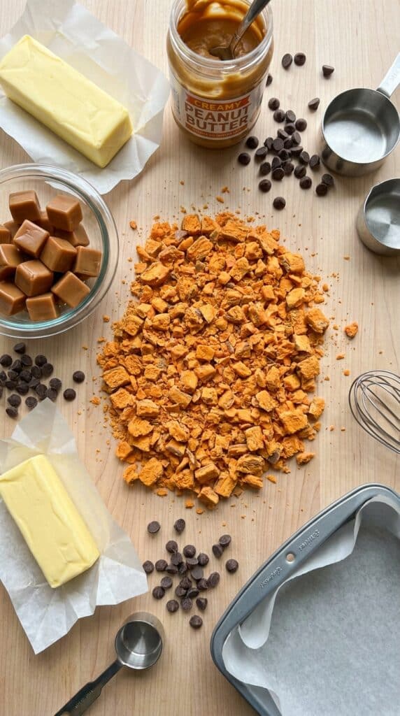 Flat lay of baking ingredients including crushed Butterfinger candy, caramels, chocolate chips, and peanut butter on a wooden surface.