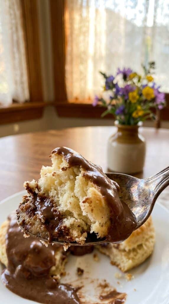 A close-up of a spoon lifting a piece of fluffy biscuit soaked in thick, dark chocolate gravy.