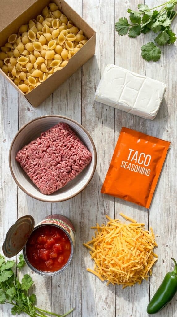 An organized layout of dry pasta, cream cheese, ground beef, taco seasoning, and canned tomatoes on a wooden table.