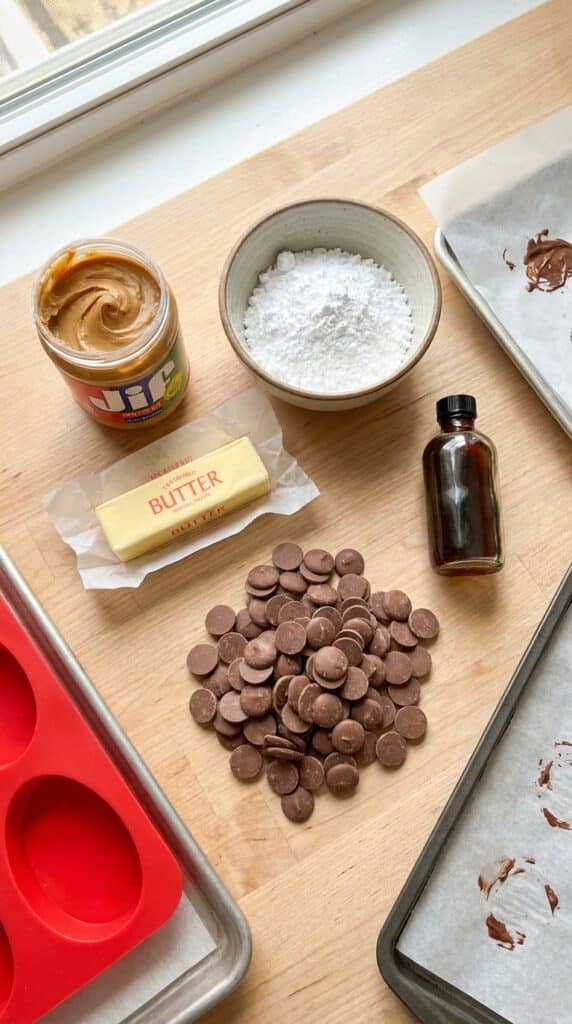 A flat lay of peanut butter, powdered sugar, butter, vanilla, and chocolate wafers on a wooden surface.