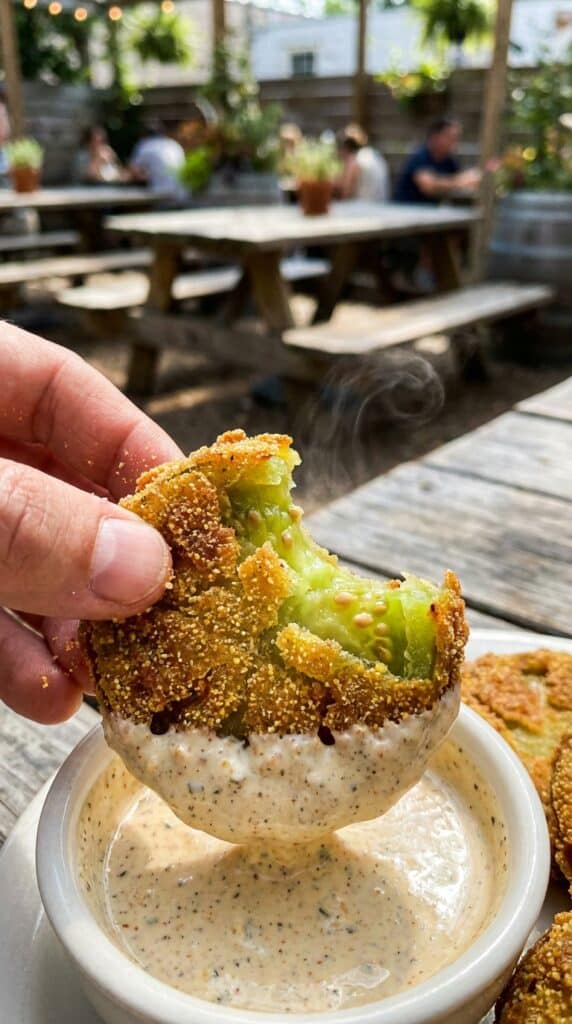 A close-up of a hand dipping a hot, crispy fried green tomato into a creamy orange sauce, showing the green tomato center.