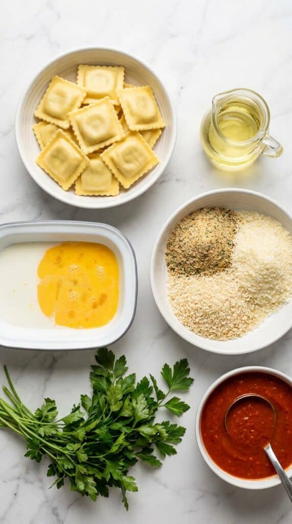 A flat lay of cheese ravioli, whisked eggs, breadcrumbs, parmesan, and marinara sauce on a marble counter.