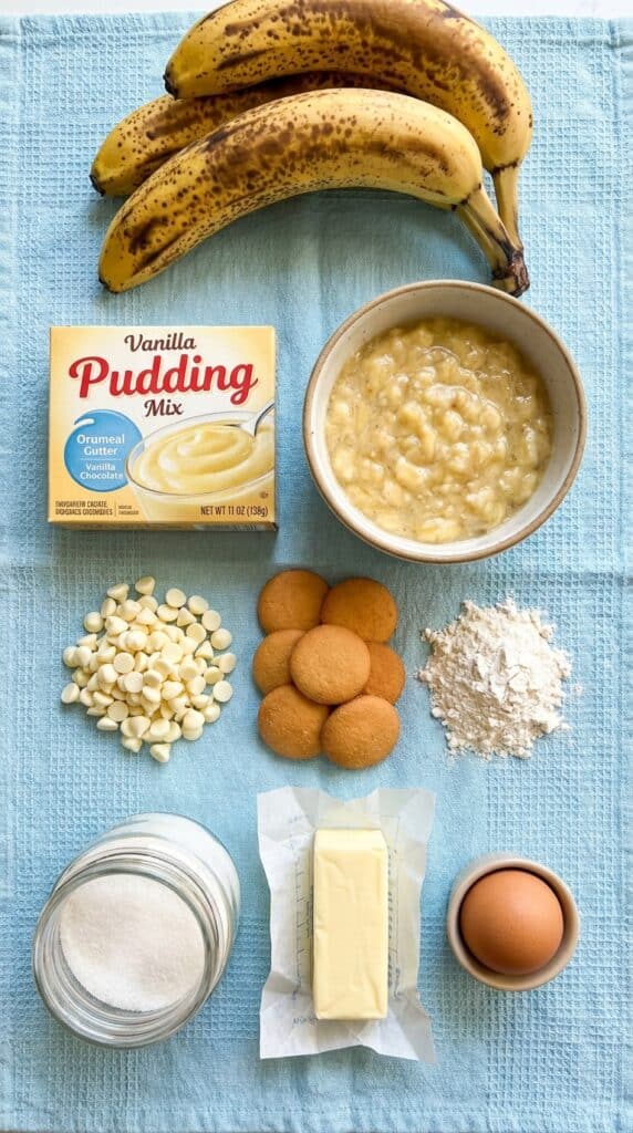 An overhead flat lay of baking ingredients: vanilla pudding mix, mashed bananas, white chocolate chips, and vanilla wafers.