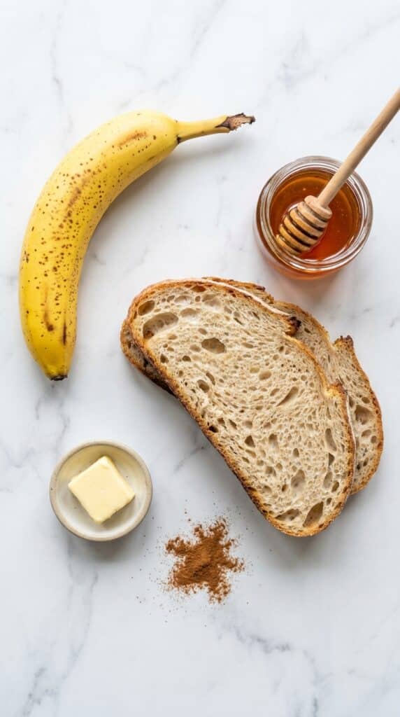 A flat lay showing a ripe banana, sourdough bread slices, a jar of honey with a dipper, and a small dish of cinnamon.