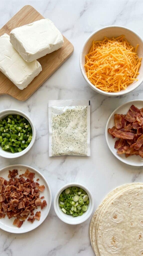 A flat lay of cream cheese, shredded cheddar, diced jalapenos, bacon bits, and tortillas on a marble counter.