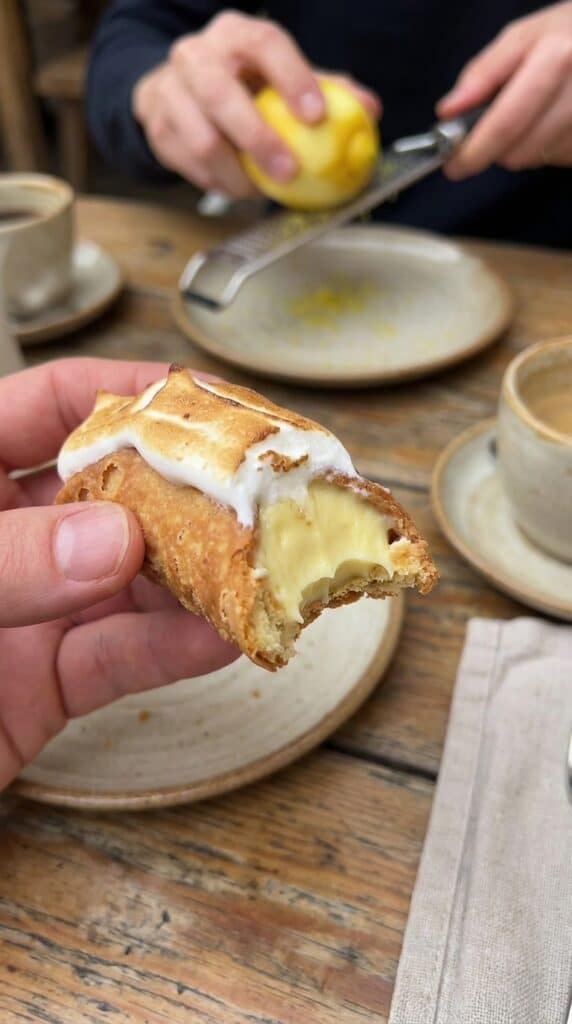 A close-up of a hand holding a lemon cannoli with a bite taken out, showing the creamy yellow filling.