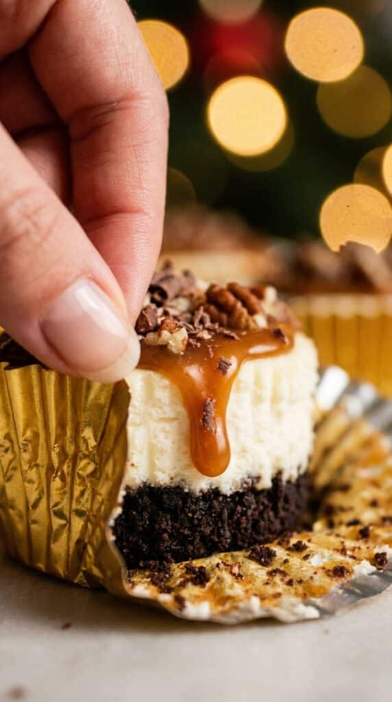 A hand peeling the wrapper off a mini turtle cheesecake, revealing the chocolate crust and creamy center.
