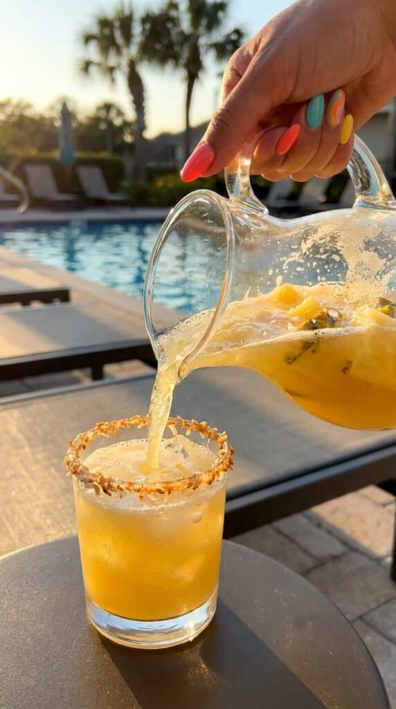Close-up of sparkling tropical sangria being poured into a glass with a toasted coconut rim by a pool.