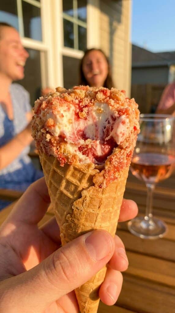 A close-up of a hand holding a strawberry crunch cheesecake cone with a bite taken out, showing the cheesecake filling and strawberry swirl.