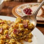 A close-up of a spoon lifting a serving of steaming bacon cornbread stuffing, showing its moist and chunky texture.