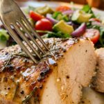 A close-up of a fork picking up a slice of juicy baked chicken, with a fresh side salad in the blurred background.