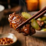 A close-up of chopsticks holding a piece of glossy black pepper chicken and celery with steam rising in a warm kitchen.