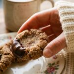 A close-up of a hand picking up a dark chocolate thumbprint cookie with a smooth ganache center, next to a cup of coffee.