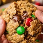 A close-up shot of a hand holding a large, thick Monster Cookie loaded with chocolate chips and holiday M&Ms.