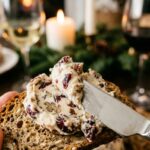 A close-up of a cheese knife spreading a creamy cranberry pecan cheese mixture onto a cracker.