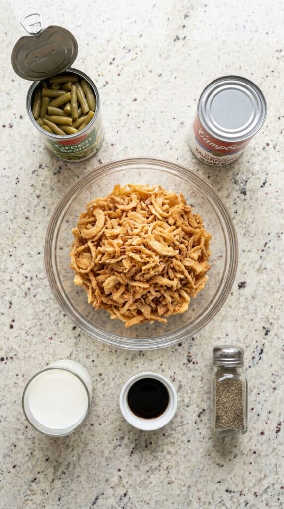 A flat lay display of canned green beans, cream of mushroom soup, milk, soy sauce, and a bowl of crispy fried onions.