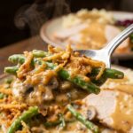 A silver spoon scooping a serving of creamy green bean casserole with crispy onions and rising steam.