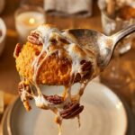 A close-up of a spoon scooping out sweet potato casserole, showing the gooey marshmallow pull and creamy orange interior.