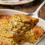 A close-up of a silver spoon scooping moist, herb-filled cornbread dressing from a baking dish, with steam rising.