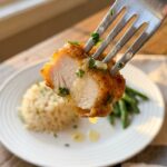 A close-up of a fork holding a juicy, golden-brown garlic butter chicken bite, with rice and green beans blurred in the background.