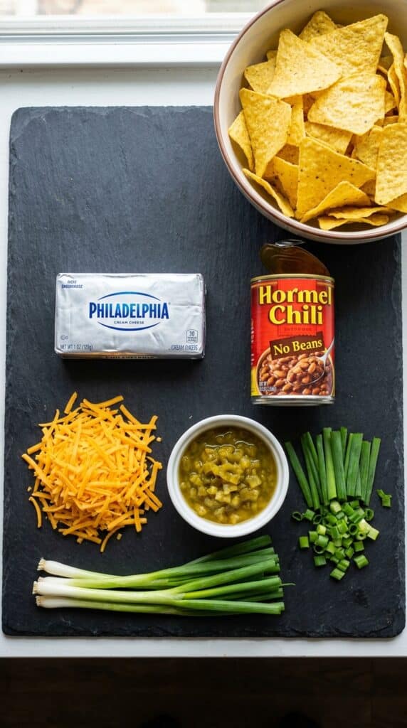 A flat lay showing cream cheese, canned chili, shredded cheddar, green chiles, scallions, and tortilla chips on a dark slate board.