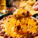 A close-up of a tortilla chip scooping up layers of cream cheese, chili, and stringy melted cheddar from a hot skillet.