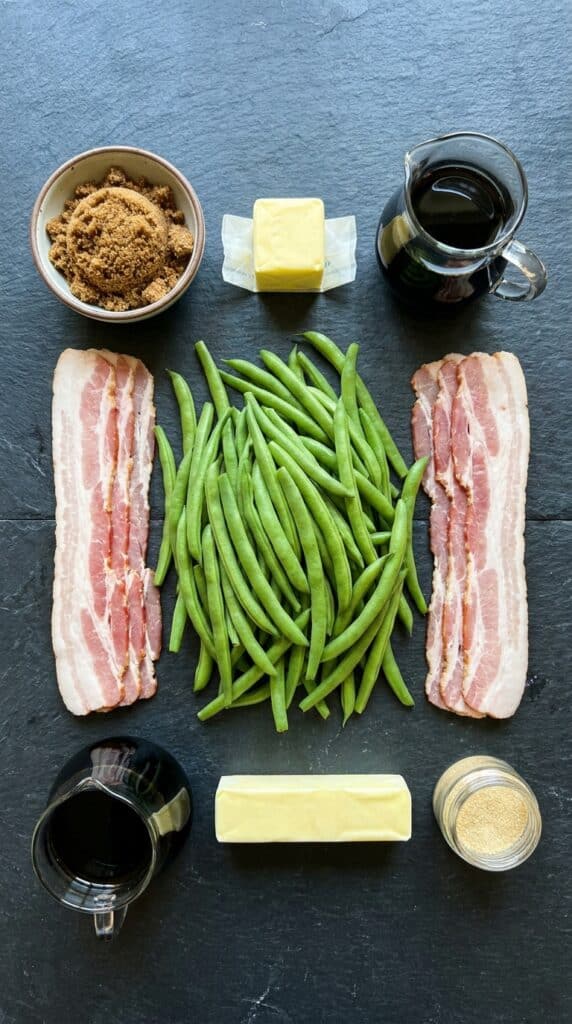 A flat lay showing fresh green beans, raw bacon, brown sugar, butter, soy sauce, and garlic powder on a dark slate board.