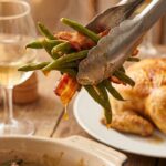 A close-up of tongs lifting sticky, glazed green beans and bacon from a dish, with roasted chicken blurred in the background.