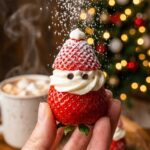 A close-up of a hand holding a single strawberry Santa treat with powdered sugar falling like snow, with a Christmas tree in the background.