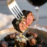 A close-up of a fork holding a single steak bite with a pink medium-rare center, dripping with garlic butter.
