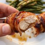 A close-up of a hand holding a sticky, bacon-wrapped chicken tender with a bite taken out, showing the juicy chicken inside.