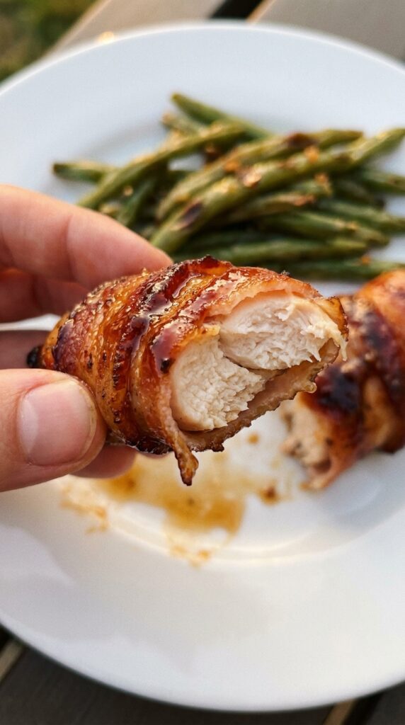 A close-up of a hand holding a sticky, bacon-wrapped chicken tender with a bite taken out, showing the juicy chicken inside.