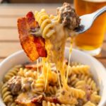 A close-up of a fork twirling cheesy pasta with a piece of bacon and ground beef, with a bowl of pasta in the blurred background.