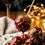 A close-up of a toothpick lifting a glossy cranberry-glazed meatball from a platter, with holiday lights in the background.