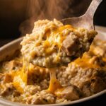 A dramatic close-up of a serving spoon lifting a steaming, creamy scoop of chicken and rice casserole with a gooey cheese pull.