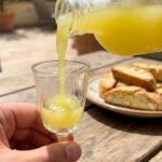 A close-up action shot of thick, bright yellow limoncello being poured into an icy cordial glass with biscotti in the background.
