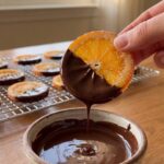 A close-up of a hand dipping a sugar-coated candied orange slice into a bowl of melted dark chocolate.
