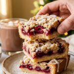 A close-up of a hand holding a sliced cherry bar showing the thick cherry layer and icing, with a Christmas tree in the background.
