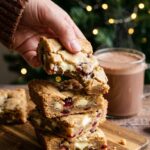 A close-up hand picking up a soft, chewy Christmas Cookie Bar, showing the soft interior with chocolate and cranberries.