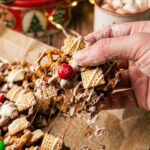A close-up of a hand picking up a large cluster of white chocolate coated cereal, pretzels, and a red M&M from parchment paper.