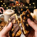 A close-up of hands breaking a piece of chocolate toffee bark in half, showing the crisp, brittle texture of the caramel and cracker.