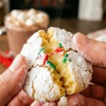 A close-up of hands breaking a powdered sugar crinkle cookie in half, revealing a soft, gooey center with holiday sprinkles.
