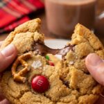 A close-up of hands breaking a thick, loaded Christmas cookie in half, showing a gooey melted chocolate center, a red M&M, and pretzel pieces.