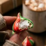A close-up of a hand holding a striped meringue kiss with a bite taken out, revealing the airy, crisp texture inside.