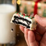 A close-up of a hand holding a white chocolate-dipped Oreo with a bite taken out, showing the dark cookie and cream center inside, with milk in the background.