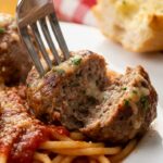A close-up of a fork cutting into a tender meatball resting on spaghetti with marinara sauce, showing a juicy interior.