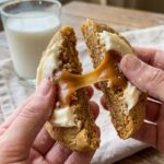 A close-up of hands breaking a large frosted caramel cookie in half, showing a soft, doughy center and a sticky caramel pull.
